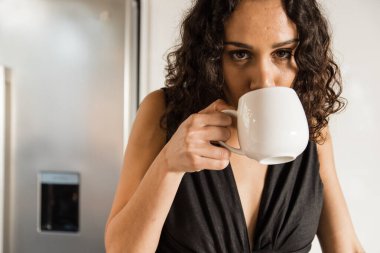 Portrait of mixed race beautiful mid adult woman with curly hair drinking fresh coffee in kitchen during morning