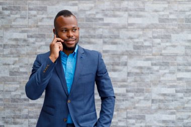 Smiling elegant male boss with hand in pocket talking over mobile phone while standing against brick wall outside office