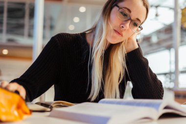 Odaklanmış, sarı saçlı, gözlük takan ve üniversitede kütüphanede masa başında ders çalışan beyaz bir kadın.