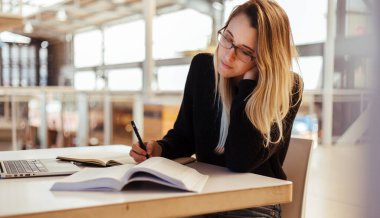 Odaklanmış, sarı saçlı, gözlük takan ve üniversitede kütüphanede masa başında ders çalışan beyaz bir kadın.