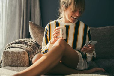 Smiling woman with blond bangs holding hot coffee and using social media over smartphone on sofa at home