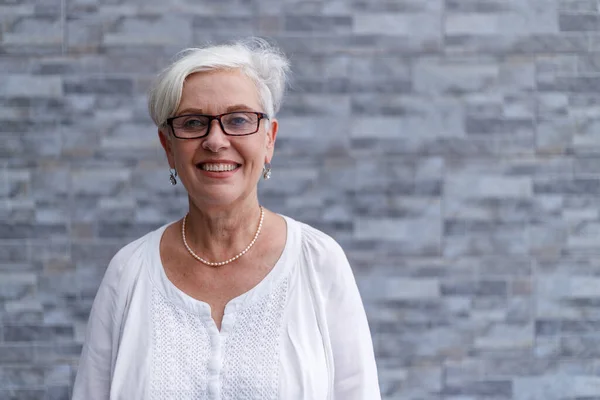 Portrait of smiling senior female executive with short hair wearing eyeglasses and standing against wall outside office