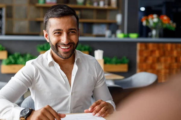 Smiling bearded young middle eastern businessman listening and looking at coworker while sitting in office cafe
