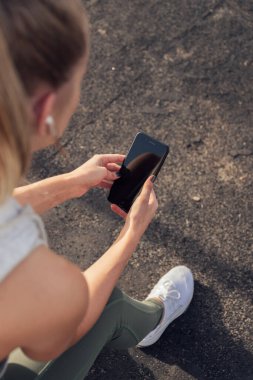 Blonde caucasian athlete resting at outdoor gym while listening to music on wireless headsets. Outdoor gym at seaside promenade.