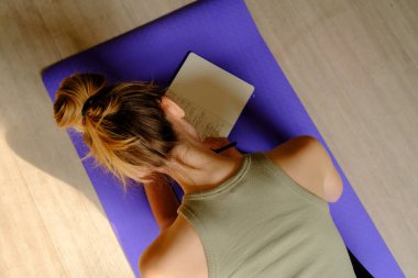 Genç beyaz kadın, evde yoga minderinin üzerinde minderin yanında duran su şişesiyle günlük tutuyor. Sağlık ve Sıhhat kavramı.