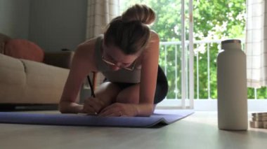 Genç beyaz kadın, evde yoga minderinin üzerinde minderin yanında duran su şişesiyle günlük tutuyor. Sağlık ve Sıhhat kavramı.