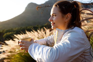 Thoughtful young woman holding coffee cup and looking away while relaxing on mountain against clear sky at sunrise