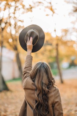 Sonbahar parkında, turuncu yapraklı ağaçların arasında şık, uzun kahverengi paltolu, modaya uygun saç stiline sahip genç bir kadın. İnsanlar, yaşam tarzı, seyahat ve tatil