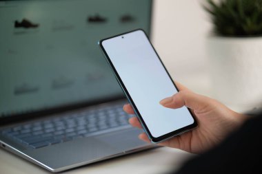 Beyaz ekranlı ve beyaz masadaki dizüstü bilgisayarlı cep telefonunu tutan ve kullanan bir kadın. 