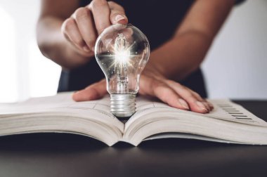 El ele tutuşup parlayan bir ampul ve açık bir kitap. Eğitim ve bilgi, iş fikri, öğrenim kavramı. İnternette öğrenme, e-öğrenme veya evde öğrenim fikri