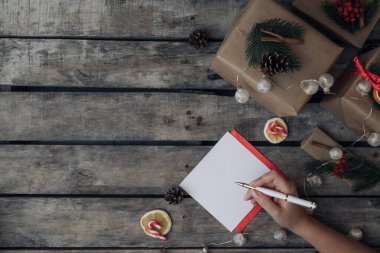 Boş noel kartına el yazısı yazan bir kadın. Noel süslemeleri, çelenkler ve Noel hediye kutularıyla taşralı ahşap bir arka plan. Üst görünüm maketi yap ve metin için alanı kopyala.
