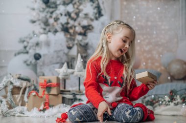 Küçük kız yılbaşı hediyesini açıyor, Noel hediyesi kutusuna bakıyor. Noel kazağı giymiş, şömine ve Noel ağacının yanında poz veriyor..