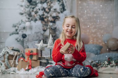Küçük kız yılbaşı hediyesini açıyor, Noel hediyesi kutusuna bakıyor. Noel kazağı giymiş, şömine ve Noel ağacının yanında poz veriyor..
