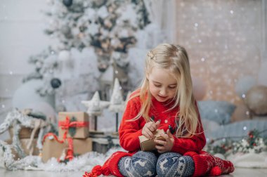 Küçük kız yılbaşı hediyesini açıyor, Noel hediyesi kutusuna bakıyor. Noel kazağı giymiş, şömine ve Noel ağacının yanında poz veriyor..