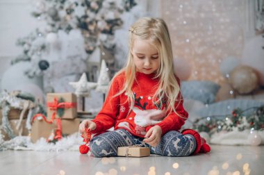Küçük kız Noel hediyesini paketliyor. Noel kazağı giyen çocuk şömine ve Noel ağacının yanında poz veriyor..