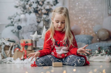 Küçük kız Noel hediyesini paketliyor. Noel kazağı giyen çocuk şömine ve Noel ağacının yanında poz veriyor..