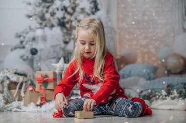 Küçük kız Noel hediyesini paketliyor. Noel kazağı giyen çocuk şömine ve Noel ağacının yanında poz veriyor..