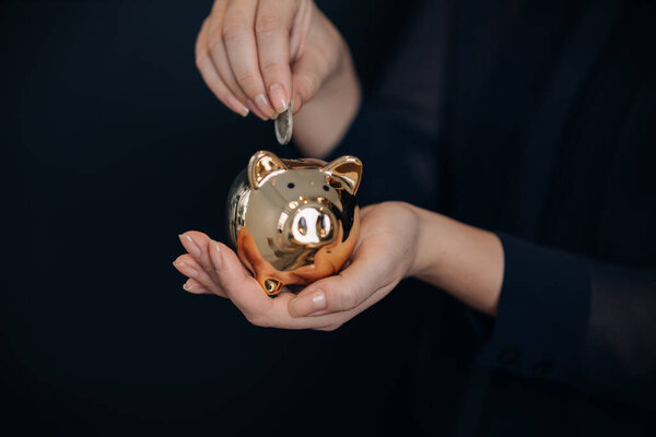 Close up of a female hand putting a coin in a golden piggy bank.Concept of saving money or savings, investment