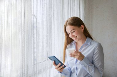 Bir kız pencerenin yanında duruyor, elinde bir fincan kahve tutuyor ve telefona bir mesaj yazıyor. Pencerenin yanında günaydın konsepti. Telefonla günaydın de.