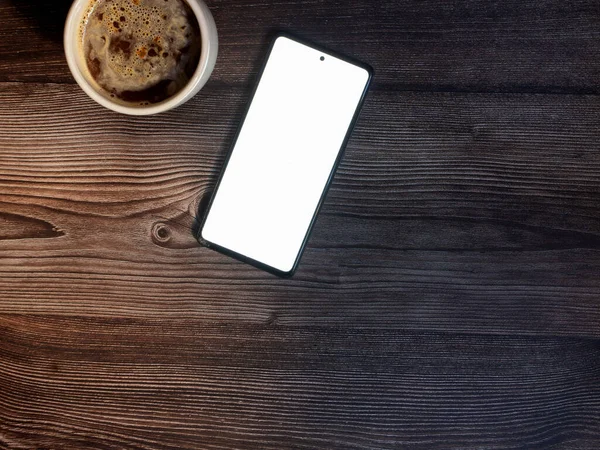 Koyu ahşap üzerinde kahve fincanı olan düz bir cep telefonu uzanıyor.