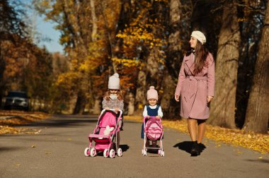 Anne ve iki kız bebek arabası sonbahar parkında yürüyor..