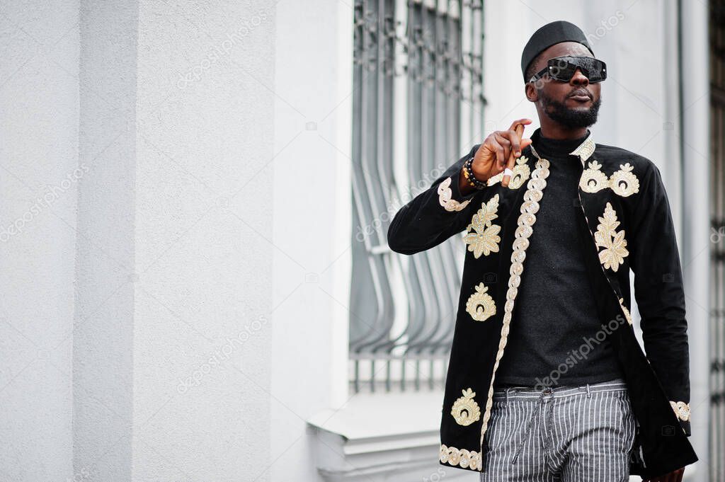 Mega stylish african man in traditional jacket pose. Fashionable black guy in hat and sunglasses with cigar in hand.