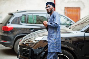 Mavi otantik kostümlü, gözlüklü ve bereli Afrikalı adam. Siyah SUV arabasının yanında şık bir Nijeryalı..