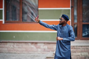 Mavi otantik kostümlü, gözlüklü ve bereli Afrikalı adam. Moda Nijeryalı adam cep telefonuyla selfie çekiyor..
