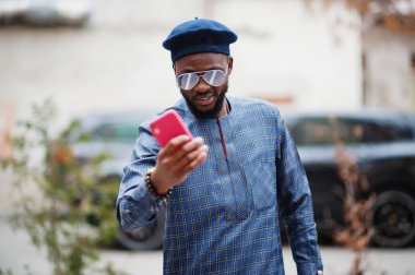 Mavi otantik kostümlü, gözlüklü ve bereli Afrikalı adam. Moda Nijeryalı adam siyah jipin yanında telefonda selfie çekiyor..