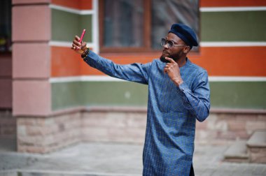 Mavi otantik kostümlü, gözlüklü ve bereli Afrikalı adam. Moda Nijeryalı adam cep telefonuyla selfie çekiyor..