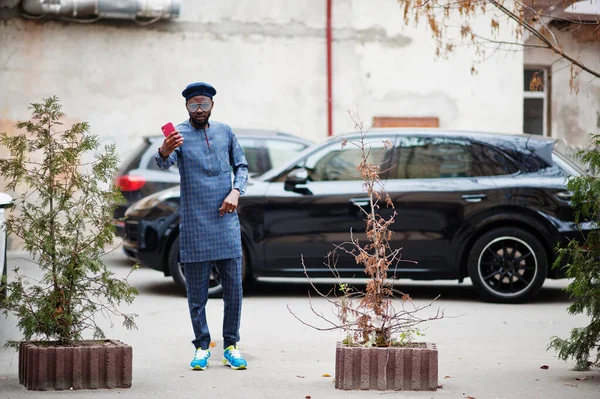 青い本物の衣装 眼鏡とベレー帽のアフリカの男 携帯電話で自慰行為をする黒のSuv車の近くのファッショナブルなナイジェリア人男性 — ストック写真