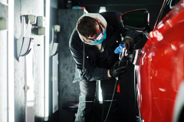 Araba temizleme konsepti. Yörünge parlatıcısı olan maskeli bir adam tamircide turuncu SUV araba cilalıyor..