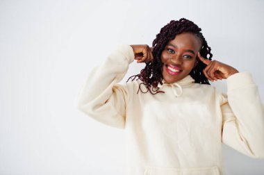 Just think about it. African woman in hoodie stand against white wall and touch fingers to temple of the head.