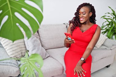 Lüks bir dairede koltukta cep telefonuyla oturan lüks kırmızı elbiseli muhteşem bir Afrikalı kadın. Güzellik, moda.