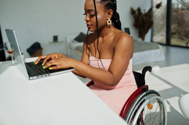 Evde dizüstü bilgisayarla çalışan engelli, tekerlekli sandalyedeki genç Afrikalı Amerikalı kadın..