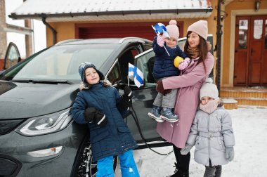 Çocuklu genç finiş anne kışın evinin bahçesinde Finlandiya bayrakları ve elektrikli araba taşıyor..