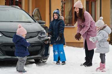 Genç anne, kışın evinin bahçesinde çocuklarla elektrikli arabaya karşı oynuyor..