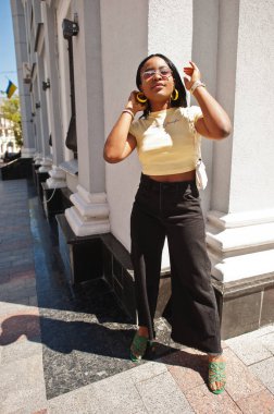 Güzel, modaya uygun, Afro-Amerikalı bir kadın sokakta poz veriyor, güneş gözlüğü takıyor, sarı bluzlu. Moda yaz fotoğrafı. Parlak renkler. Güzel manzara afro insanlar..