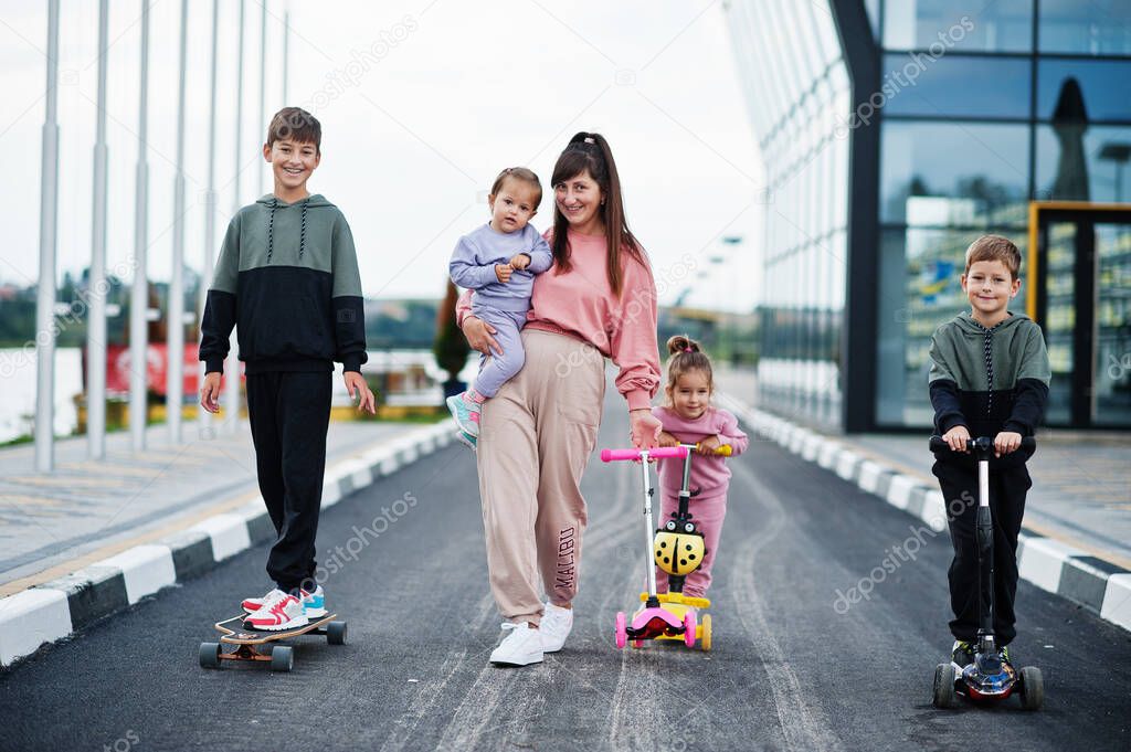 Young stylish mother with four kids outdoor. Sports family spend free time outdoors with scooters and skates.