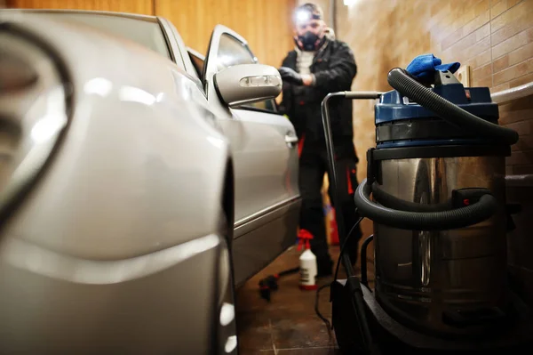 Man in uniform and respirator, worker of car wash center, cleaning car interior with hot steam cleaner. Car detailing concept.