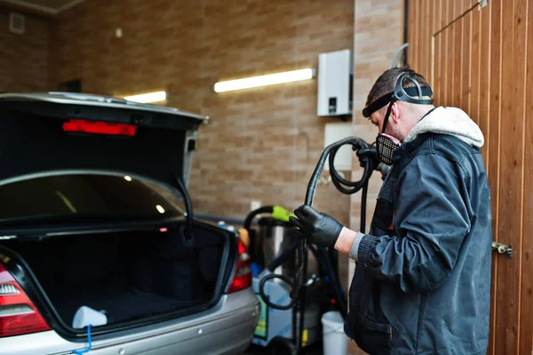 Man in uniform and respirator, worker of car wash center, cleaning car interior with hot steam cleaner. Car detailing concept.