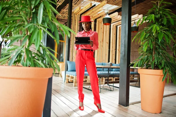 Pretty braids business african american lady bright bossy person friendly wear office red shirt, hat and trousers, hold tablet on hands.