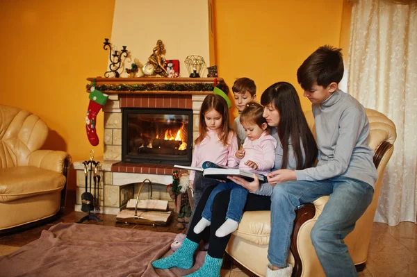 Happy young large family by a fireplace in warm living room on winter day. Mother with four kids at home read book.