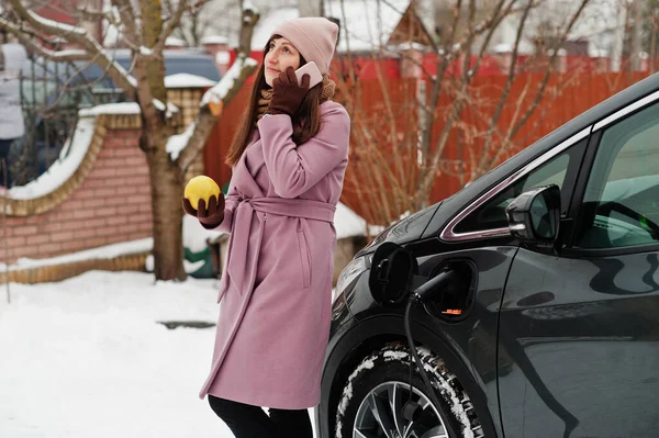 Young woman with mobile phone and citrus lemon fruit at hand charging her electric car in winter. Eco transport concept.