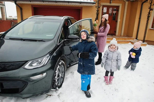 Young woman with kids hold eco bags and charging electric car in the yard of her house .