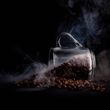 Glass, transparent mug, overturned. Hundreds of aromatic coffee beans fall out of it. Black background, smoke that looks magically fragrant. Place for ad text. Photo background for coffee drinkers.
