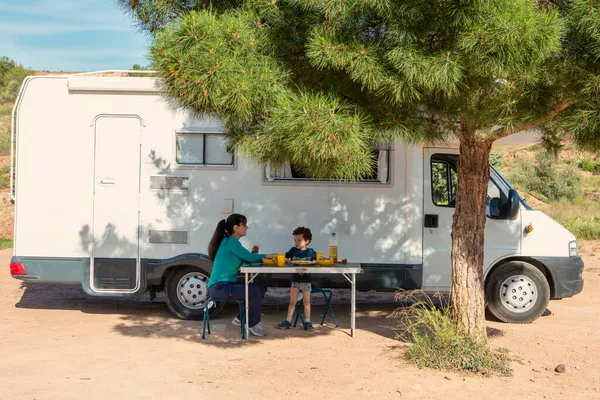 Anne ve oğlu karavanın yanındaki kamp masasında, bir ağacın gölgesinde.