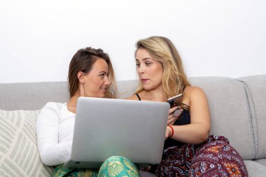 Kanepede dizüstü bilgisayara ve akıllı telefona bakan gülümseyen lezbiyen çift.