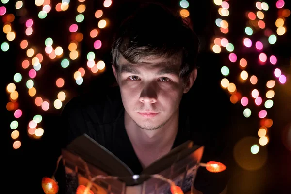Un joven se sienta con un libro en sus manos, iluminado por guirnaldas de colores en un ambiente fabuloso. — Foto de Stock