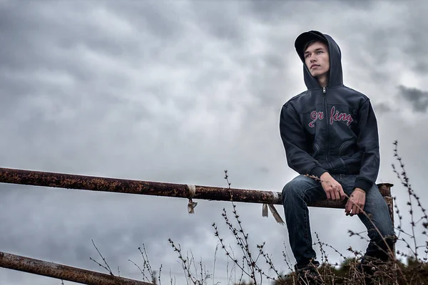 Ein junger Mann mit Kapuze geht auf einer Absperrung vor einem bewölkten grauen Himmel Stockbild